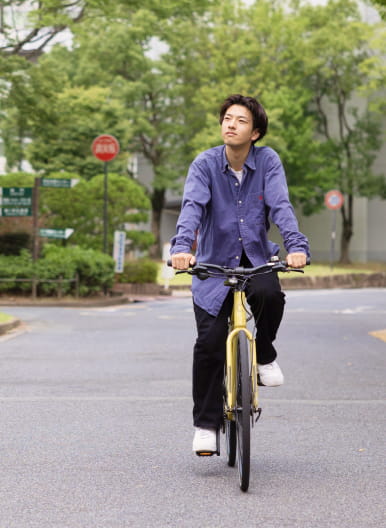 緑の木々に囲まれた道路を、青いシャツを着た男性が黄色い自転車で走っている様子