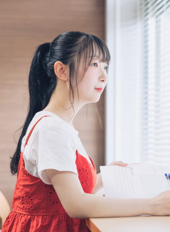 赤いワンピースを着た女性が、机に座って書類を持ちながら窓の外を見ている様子。