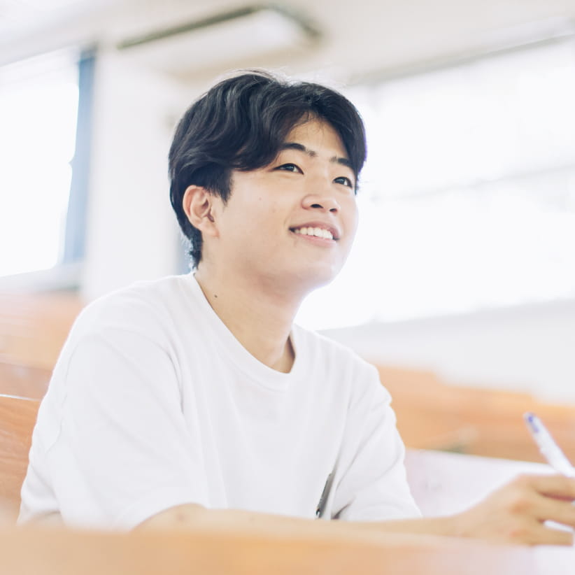 白いTシャツを着た若い男性が、教室の席に座りながら笑顔で前を見つめている様子