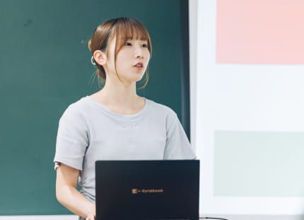 教室の黒板前で、ノートパソコンを使いながら発表をしている女性の姿