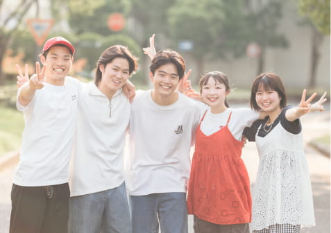 屋外で肩を組みながら並び、笑顔でピースサインをする5人の若者たち