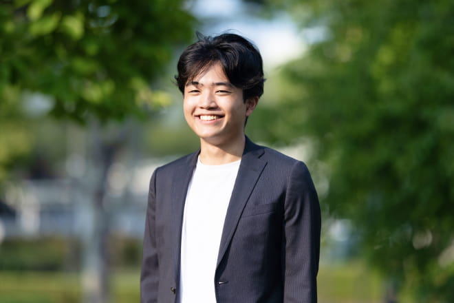 緑あふれる屋外でスーツ姿の若い男性が笑顔で立っている様子