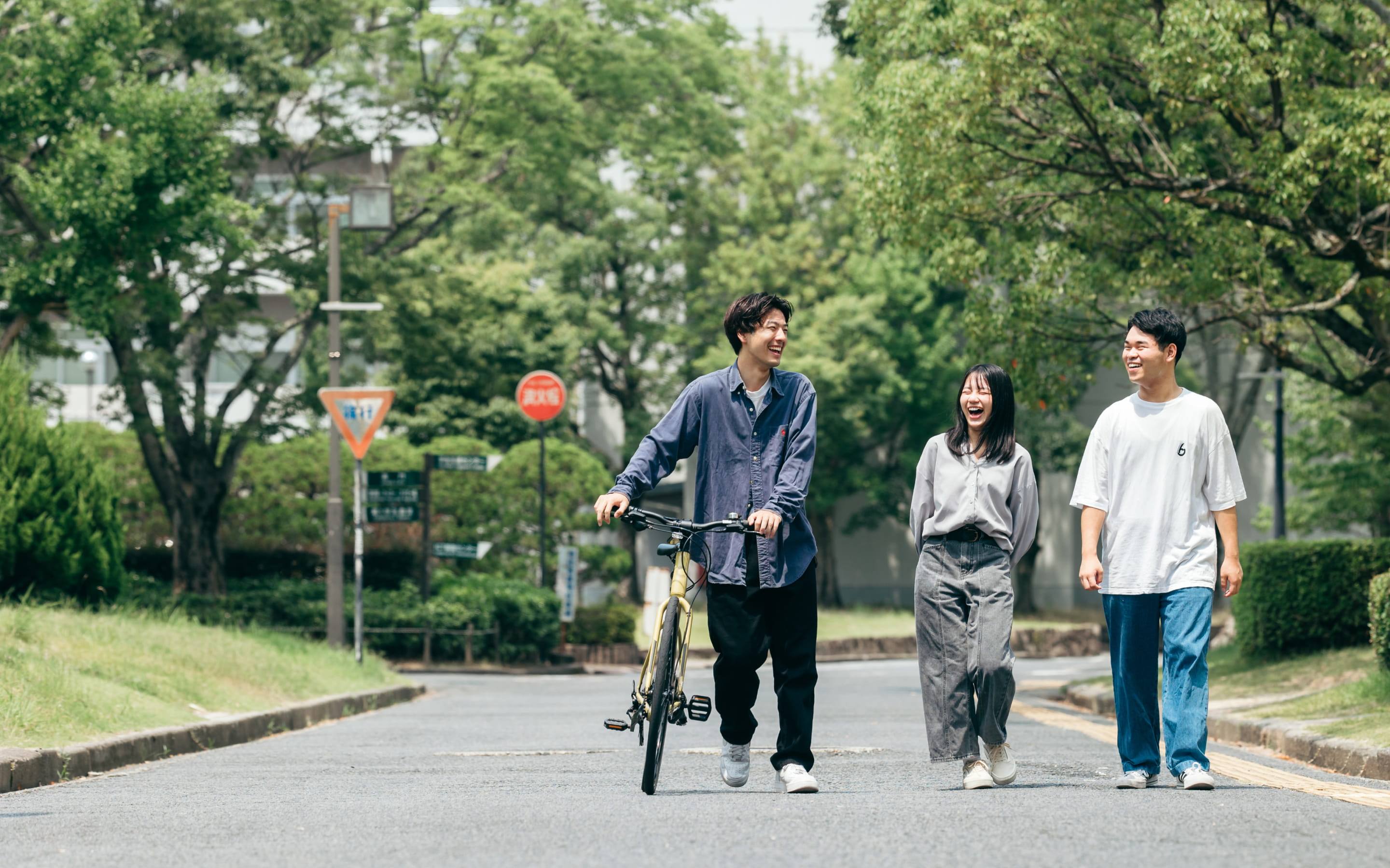 緑に囲まれた道を、笑顔で会話しながら歩く3人の大学生と自転車を押す男性の様子