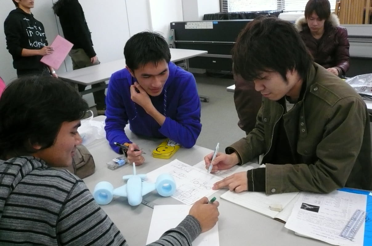 教室内で学生たちが模型飛行機を使いながら設計図やノートを広げて話し合っている様子