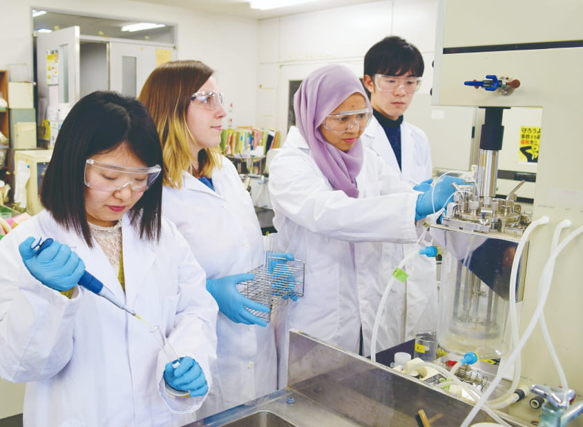 複数の学生が白衣と保護具を着用し、理化学機器を使って実験を行っている研究室の様子