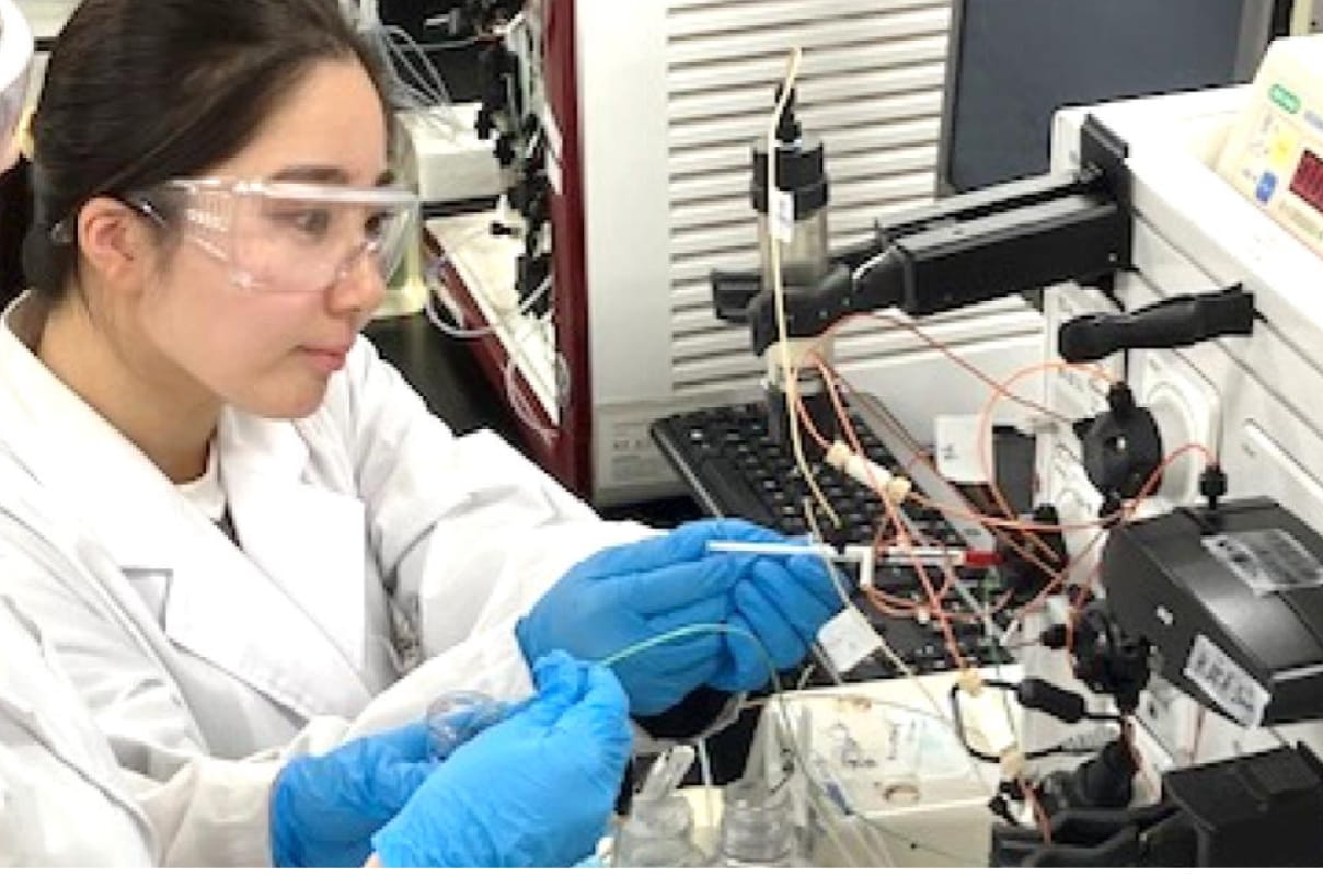 白衣とゴーグルを着用した学生が、実験装置を操作しながら測定を行っている様子
